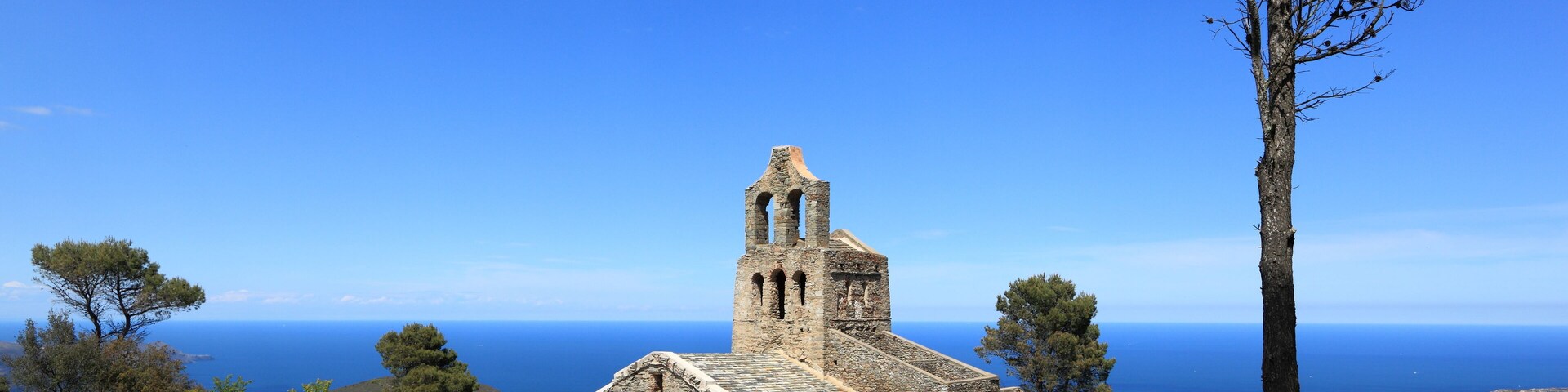 Una iglesia de piedra antigua con un campanario, rodeada de árboles y con vistas al paisaje costero.