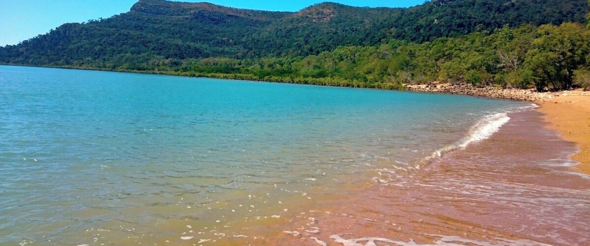 A beach with clear turquoise water, a sandy shore, and a forested mountain in the background.