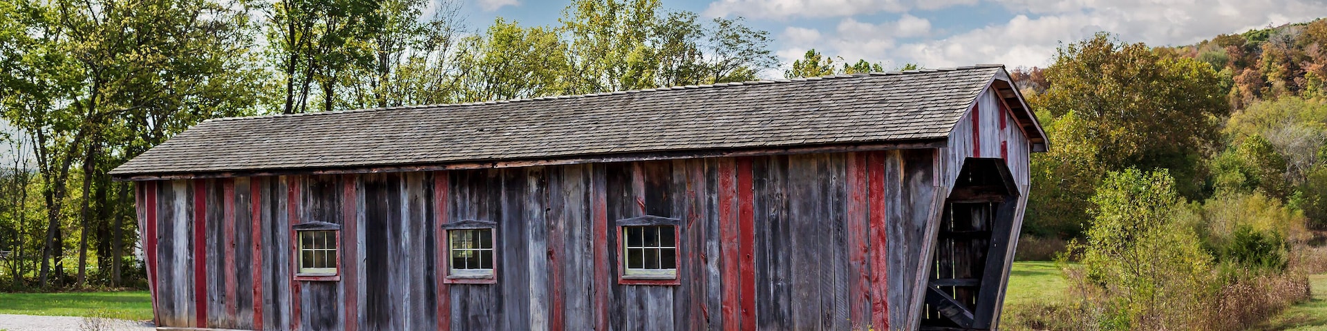 A rustic covered bridge with a wooden structure and red accents, surrounded by lush greenery and trees.