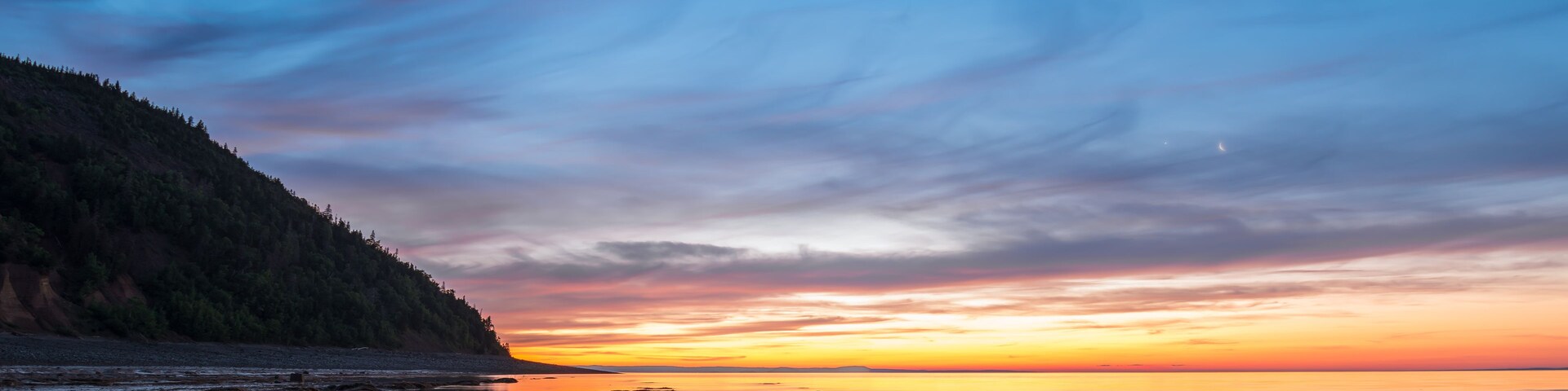 Un lago tranquilo al atardecer, con una ladera boscosa a la izquierda y un cielo despejado.