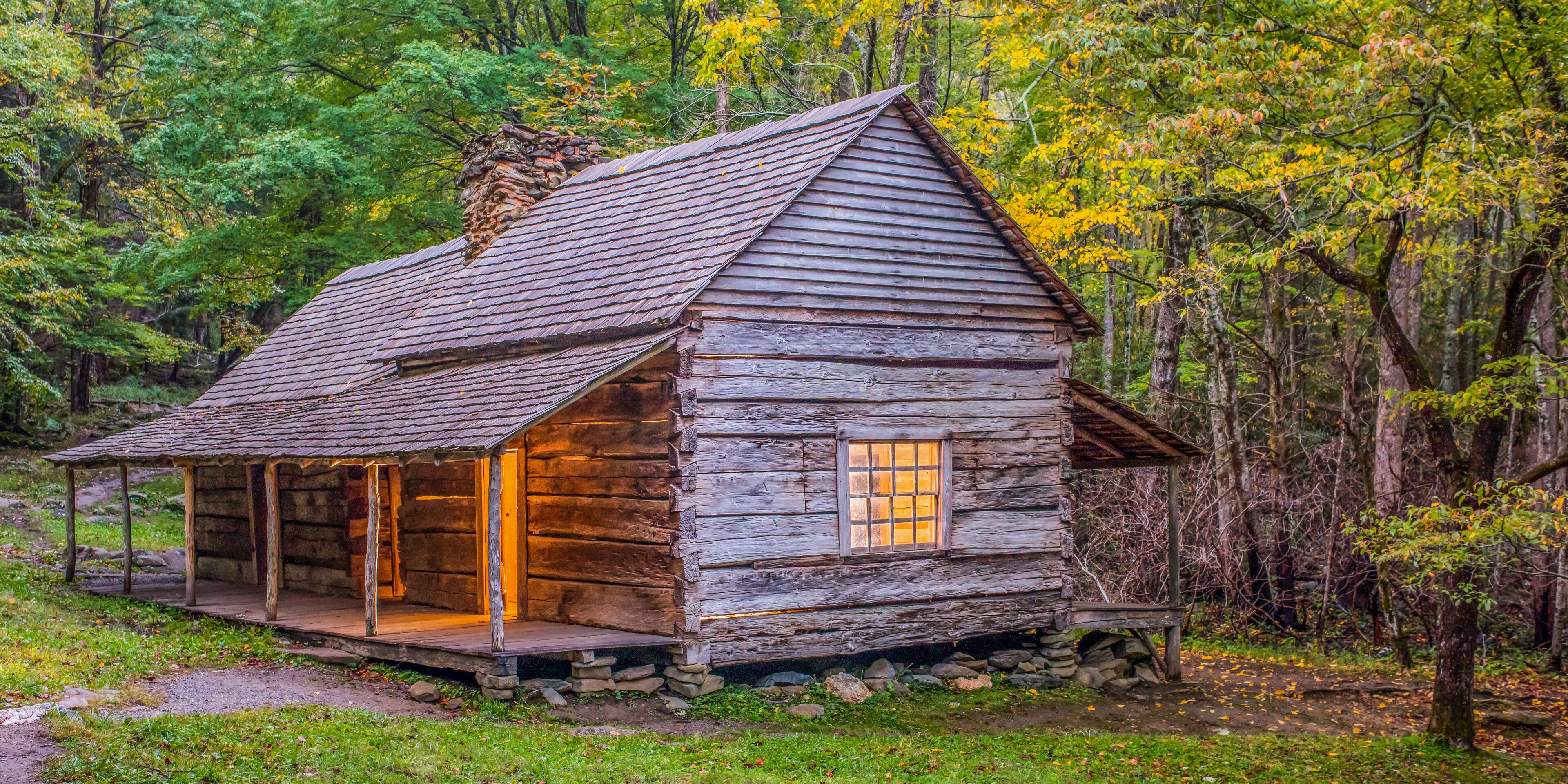 A rustic log cabin with a sloped roof, surrounded by a forest with autumn-colored foliage.
