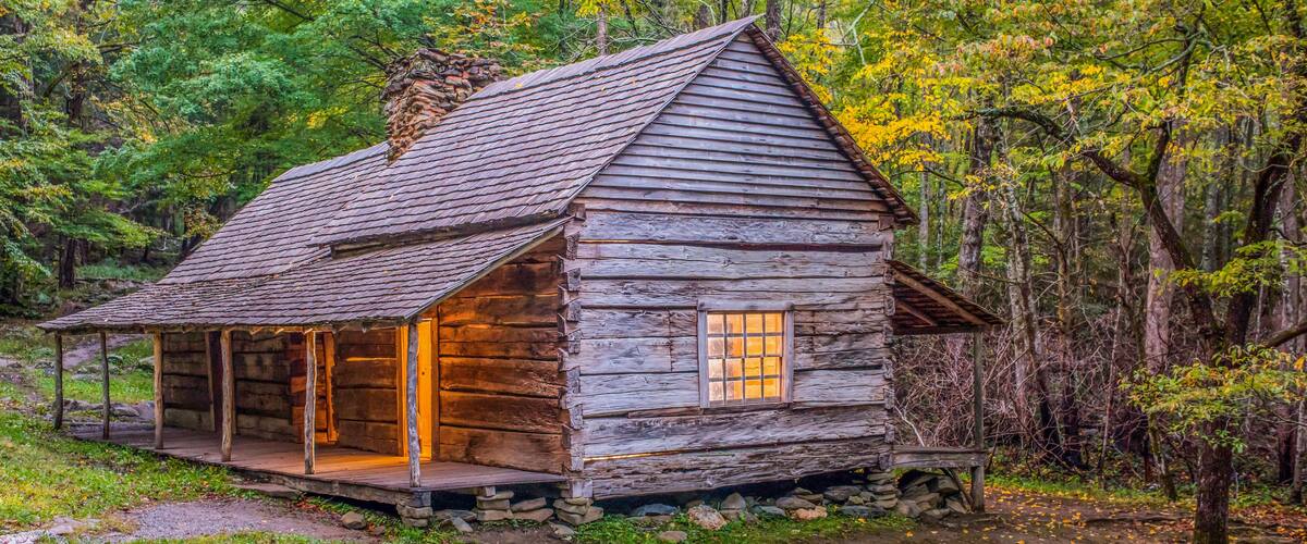 A rustic log cabin with a sloped roof, surrounded by a forest with autumn-colored foliage.