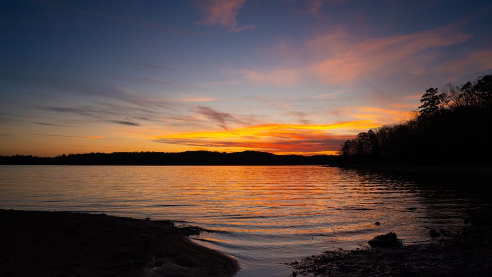 A sunset over a calm lake with a silhouette of trees on the right and a rocky shoreline in the foreground.