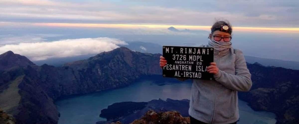 A person holding a sign with 'MT. RINJANI 3726 MDP' and 'ANGKATAN 2B PESANTREN IS AL-IRSYAD' written on it, standing on a mountain overlooking a valley with a lake.