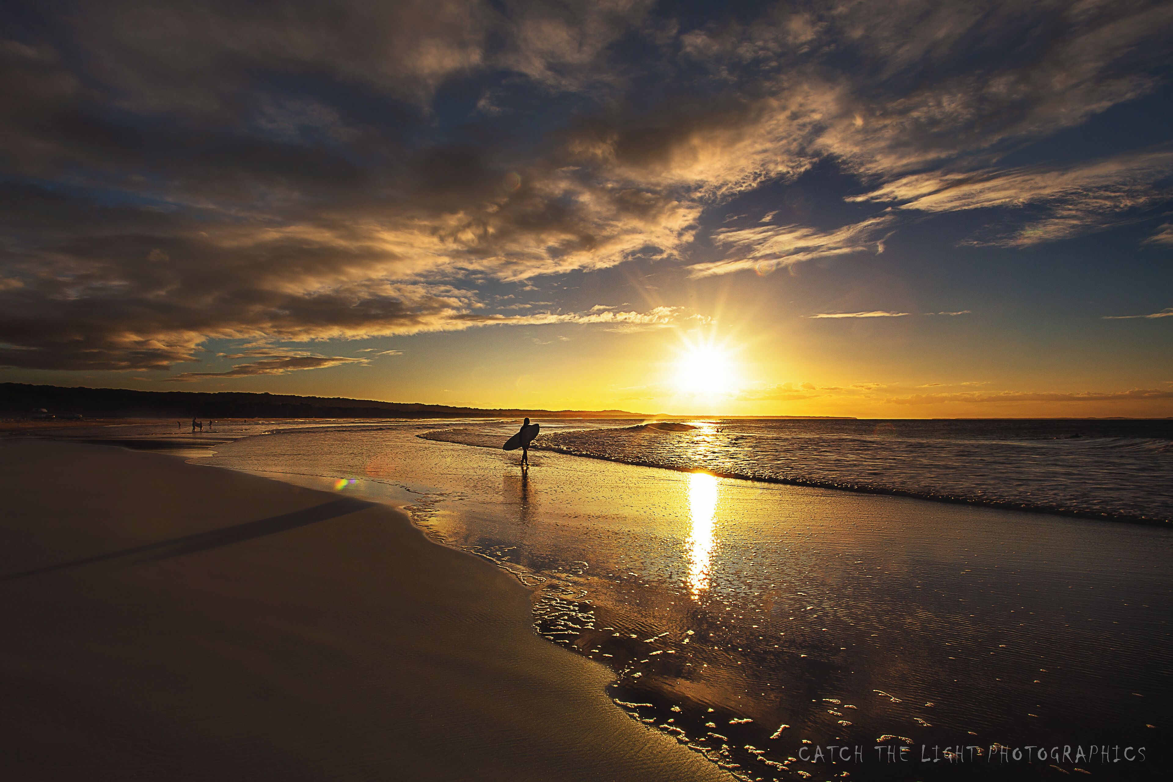 A sunset over a beach with a surfer standing on the shore.