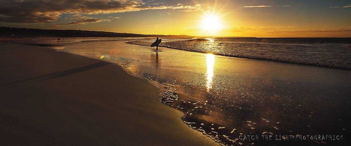 A sunset over a beach with a surfer standing on the shore.