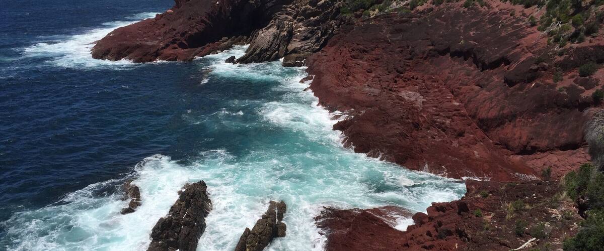 A coastal landscape with red rocks and turquoise waves crashing against them.