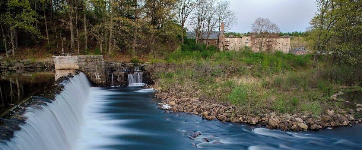 A calm river with a small waterfall, surrounded by trees and a stone bridge.