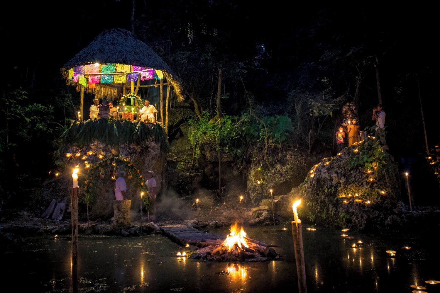 A night scene with a thatched-roof structure adorned with colorful decorations, a fire pit, and people around it.