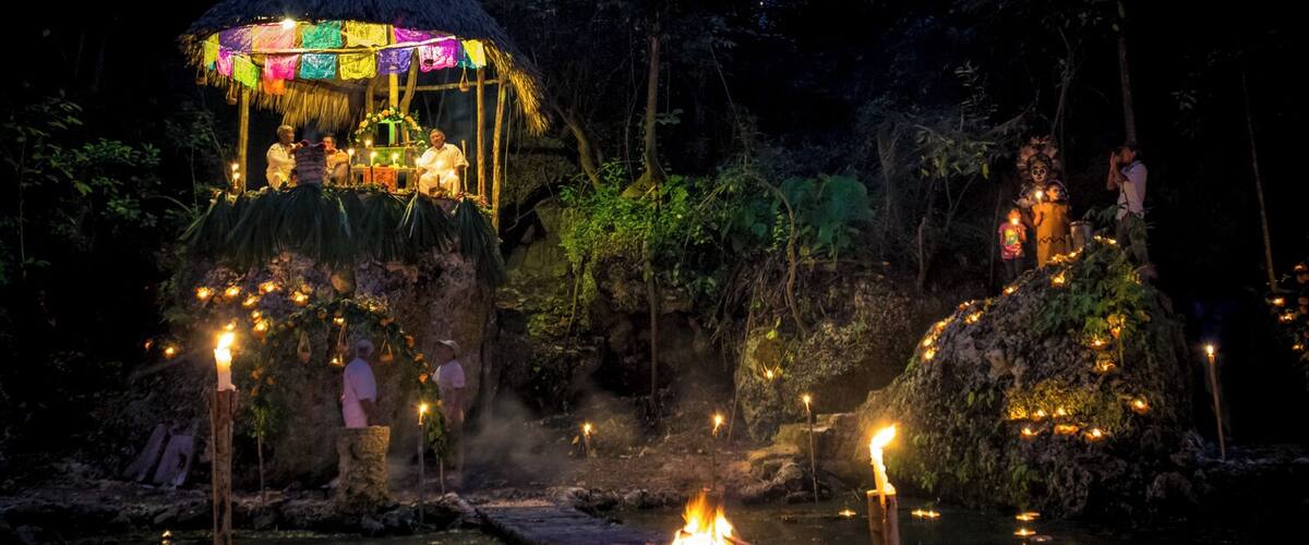 A night scene with a thatched-roof structure adorned with colorful decorations, a fire pit, and people around it.