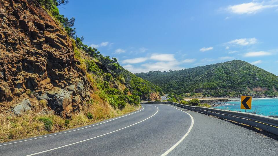 A winding coastal road with a sharp bend, rocky cliffs, and a clear blue sky.