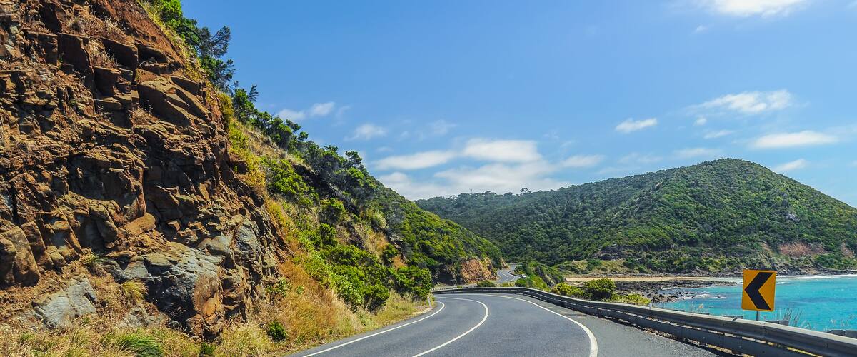 A winding coastal road with a sharp bend, rocky cliffs, and a clear blue sky.