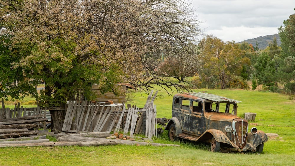 Une vieille voiture rouillée est garée dans un champ herbeux, avec une clôture en bois et un arbre en arrière-plan.