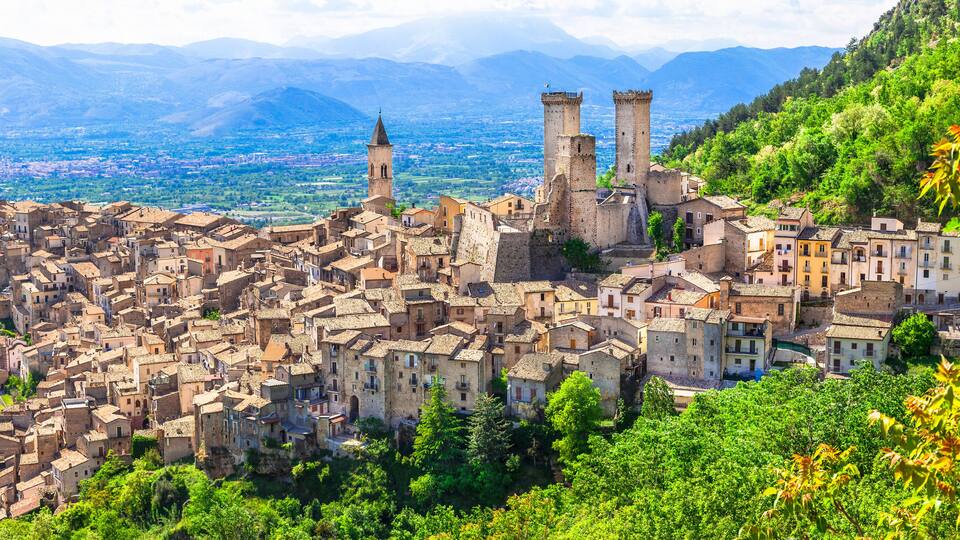 A historic town with medieval architecture, including a castle with two towers, surrounded by lush greenery and mountains in the background.
