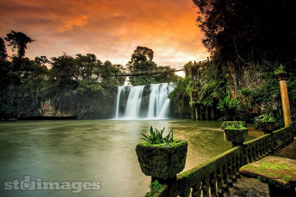 A waterfall surrounded by lush greenery and a stone bridge.