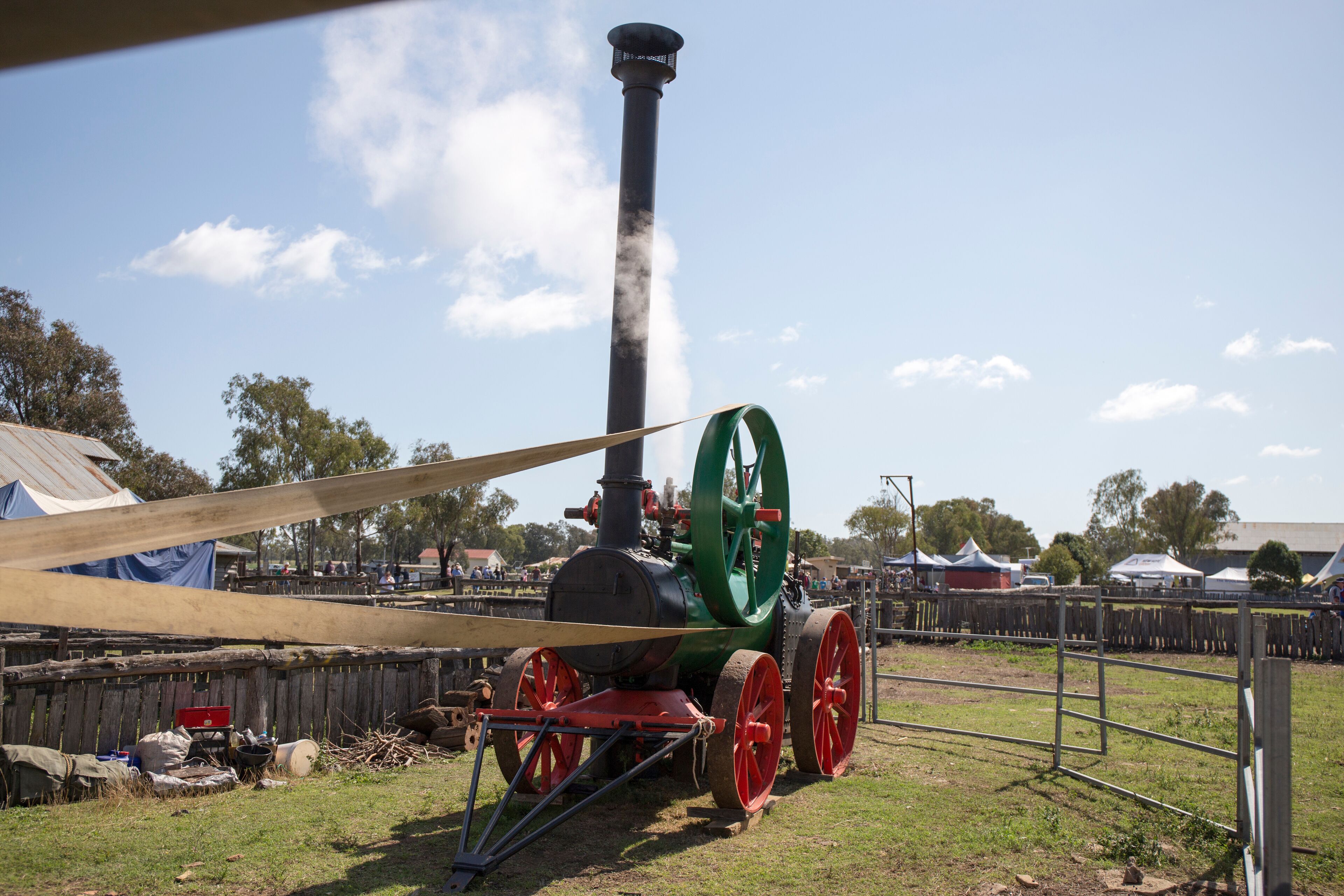 A steam locomotive with red wheels and a green front, emitting steam, parked on grass with tents and a wooden fence in the background.