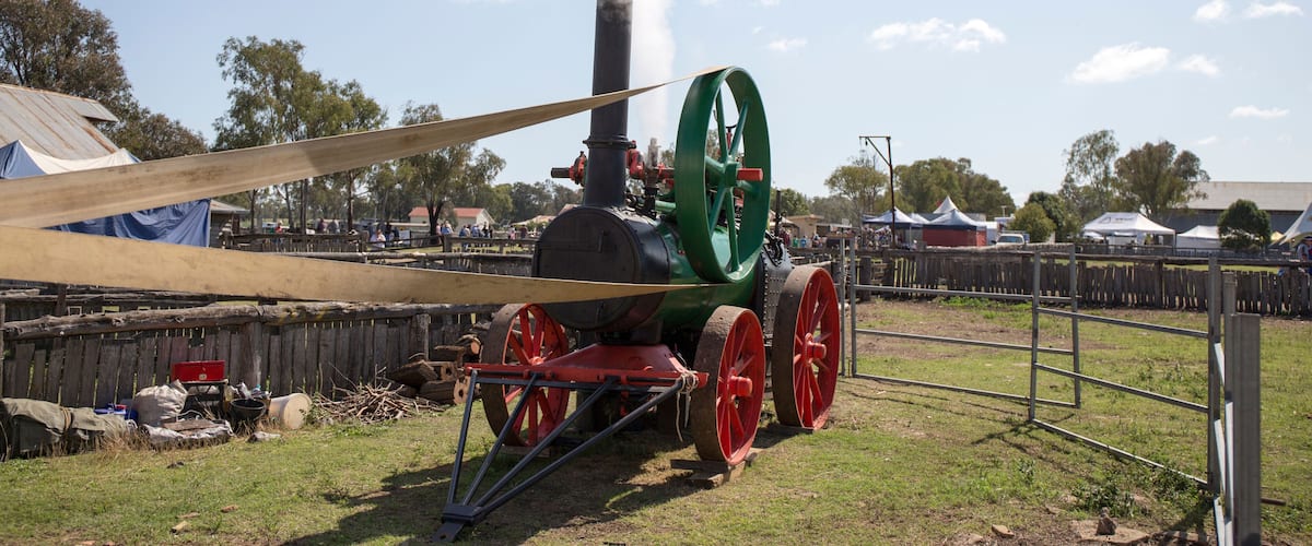 A steam locomotive with red wheels and a green front, emitting steam, parked on grass with tents and a wooden fence in the background.