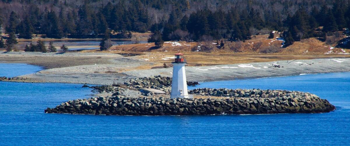 A lighthouse on a rocky island surrounded by a calm blue lake, with a forested hill in the background.