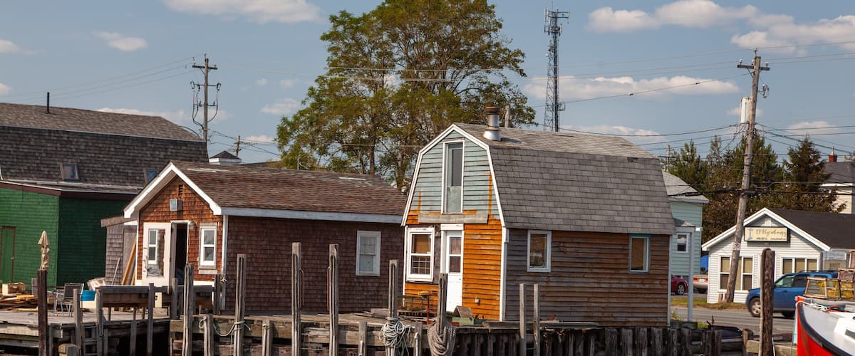 A wooden house with a steep roof, docked at a pier, surrounded by other buildings and a tree.