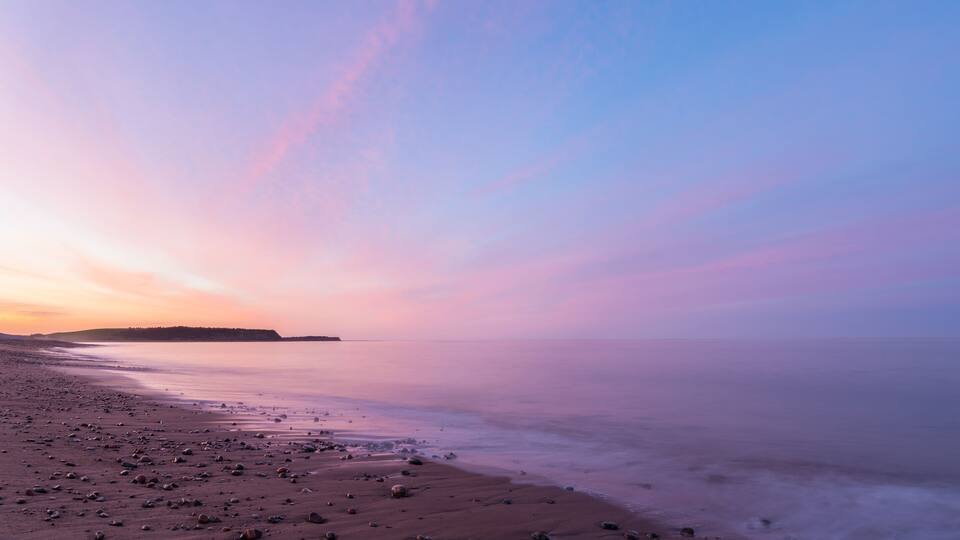 A beach with a calm sea, pebbles, and a distant island during sunset.