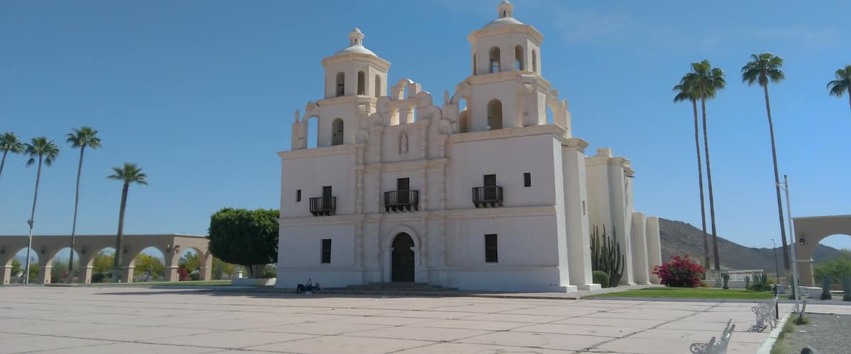 A white colonial-style church with two bell towers and arched windows, surrounded by palm trees and a clear blue sky.