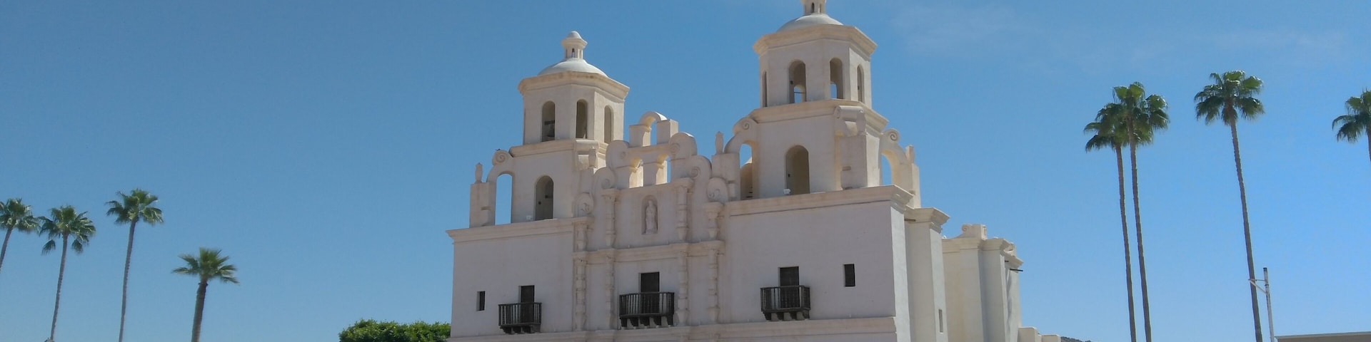 A white colonial-style church with two bell towers and arched windows, surrounded by palm trees and a clear blue sky.