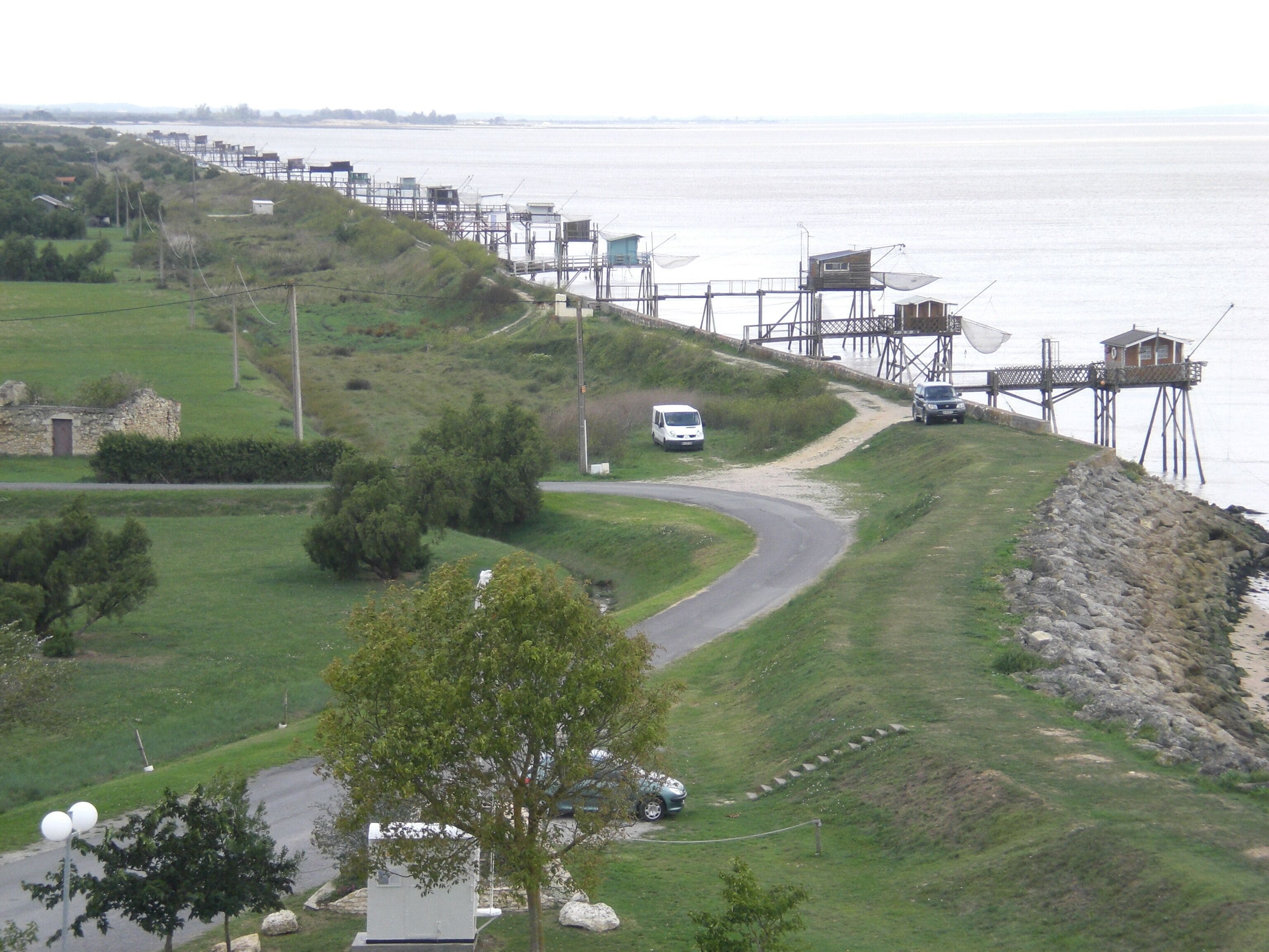 A coastal area with a road curving along the shore, a white van parked on the side, and a fishing pier extending into the water.