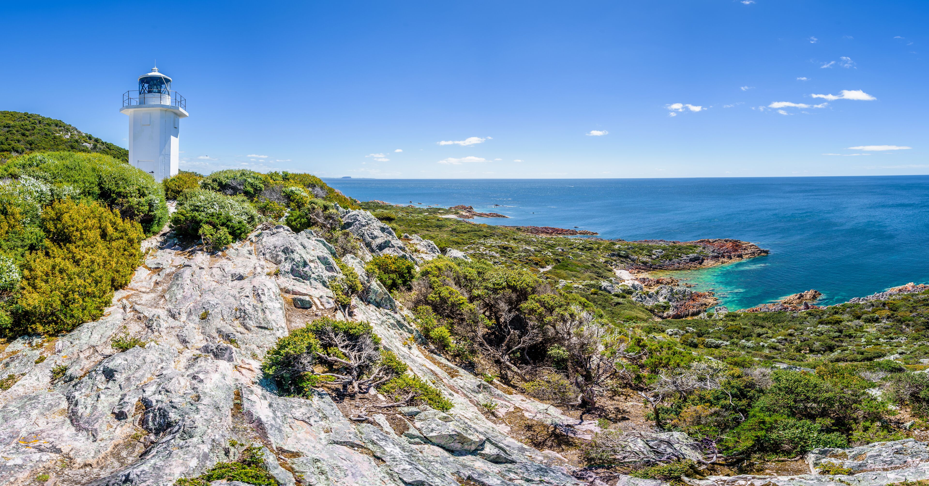 Stunning view from bright white light station to deep blue sea ocean bay turquoise water with orange red rocks at shore coast on warm sunny clear sky day, Rocky Cape National Park, Tasmania, Australia