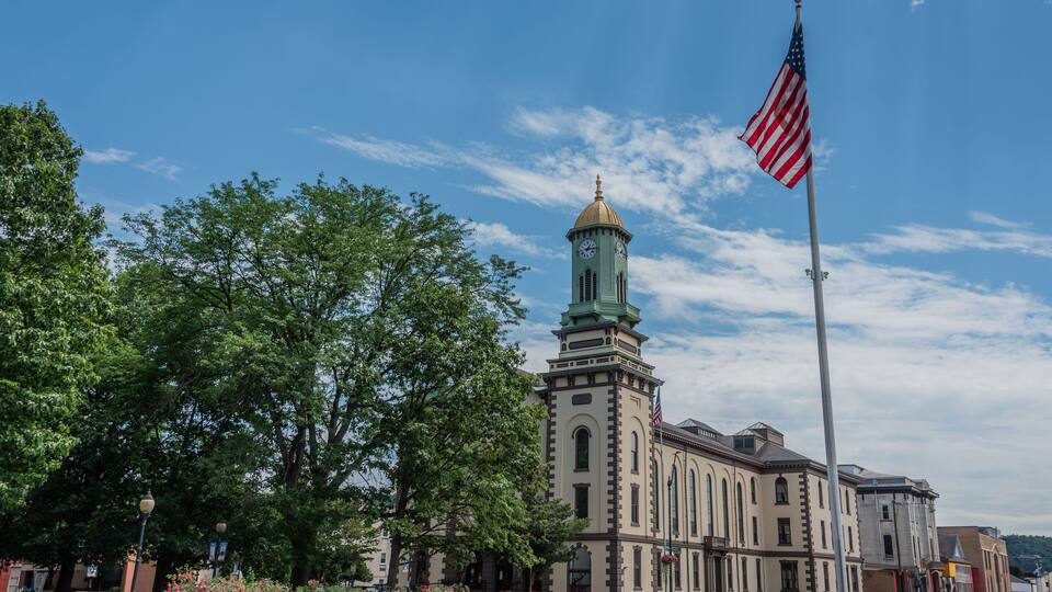 A historic building with a clock tower, surrounded by lush green trees and a flagpole with an American flag.