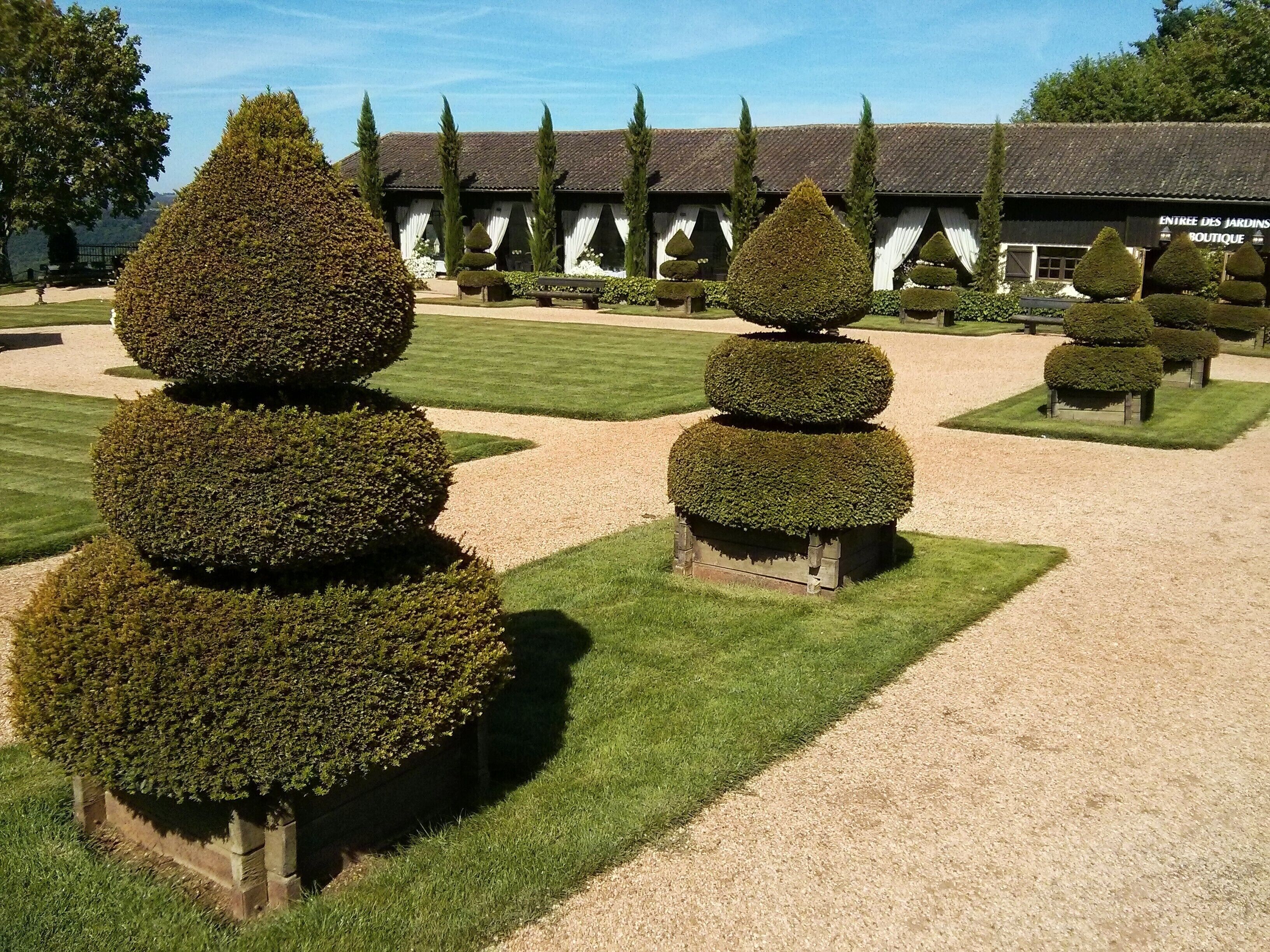 A formal garden with geometrically shaped topiary trees and a building labeled "ENTREE DES JARDINS BOUTIQUE" in the background.