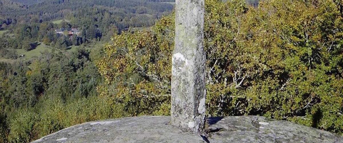 A weathered stone cross on a hilltop with a view of a forested valley below.