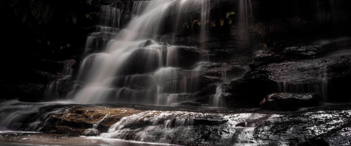 A multi-tiered waterfall cascading over dark rocks in a forested area.