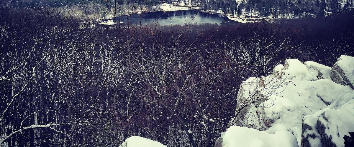 A snow-covered landscape with a lake, forested hills, and rocky outcrops.