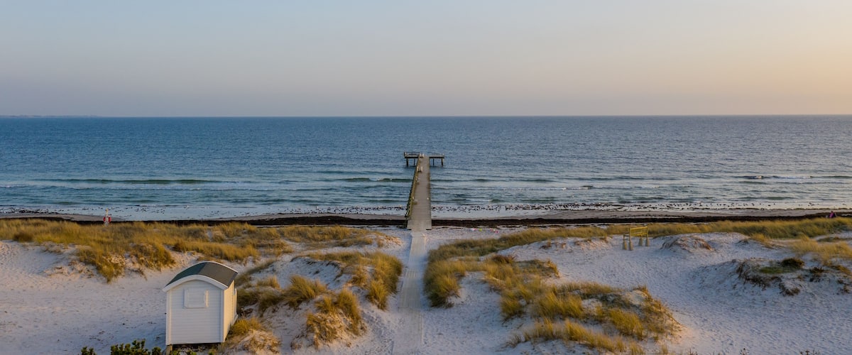 A beach with a wooden pier extending into the ocean, a small white building, and sand dunes.