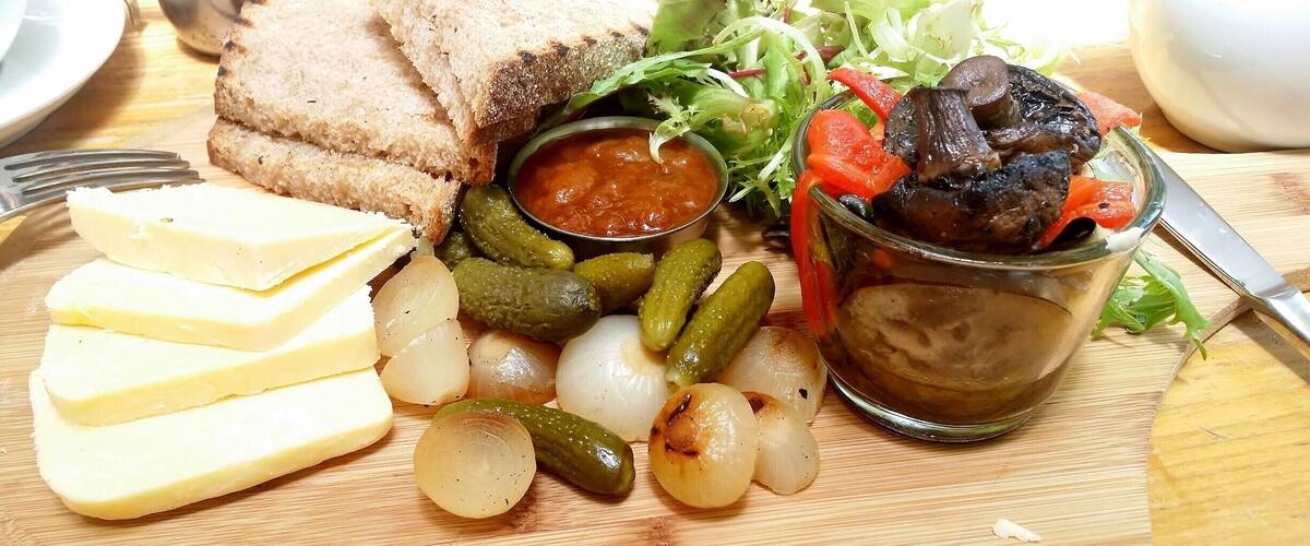 A wooden table with a variety of foods including bread, cheese, pickles, onions, a salad, and a bowl of sauce.