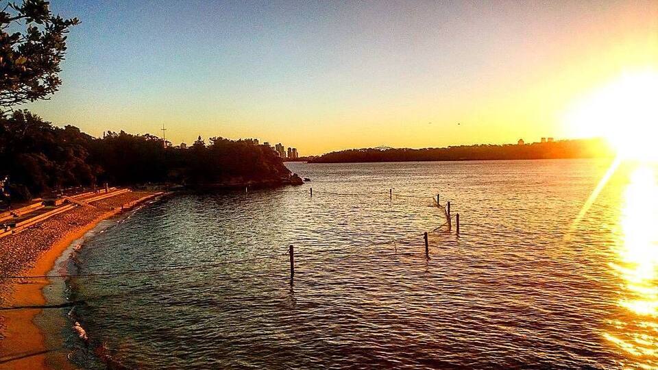 A sunset over a calm river with a beach on the left and a city skyline in the distance.
