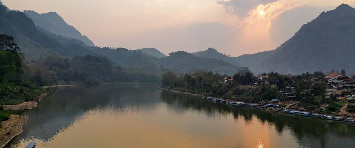 A calm river with mountains in the background and a small settlement along the riverbank.