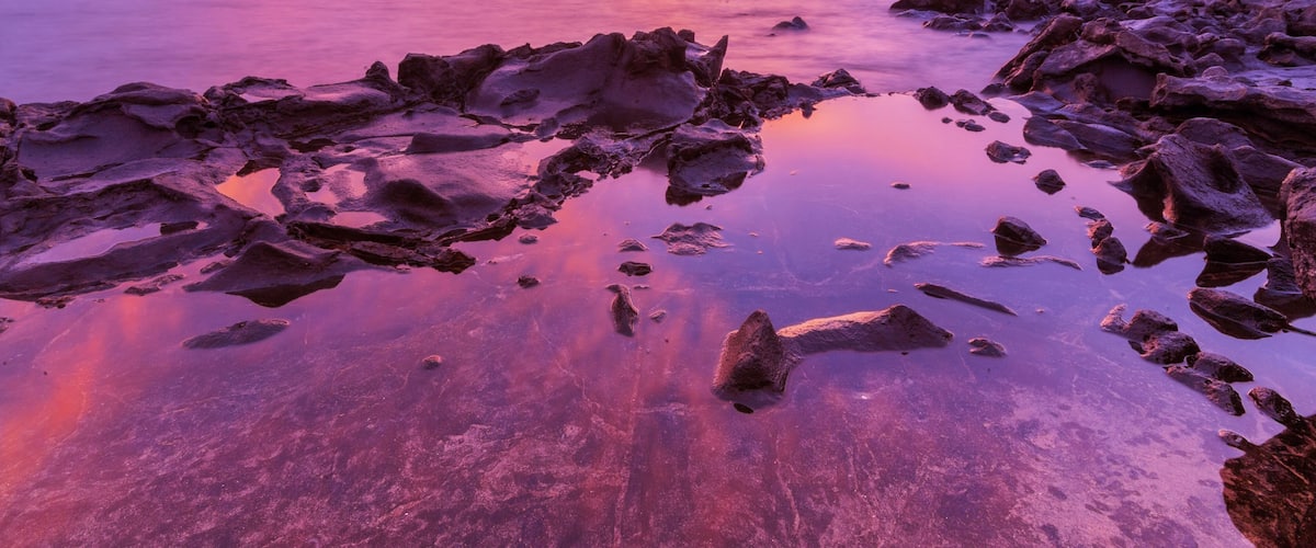 A rocky shoreline at sunset with vibrant purple and pink hues in the sky.