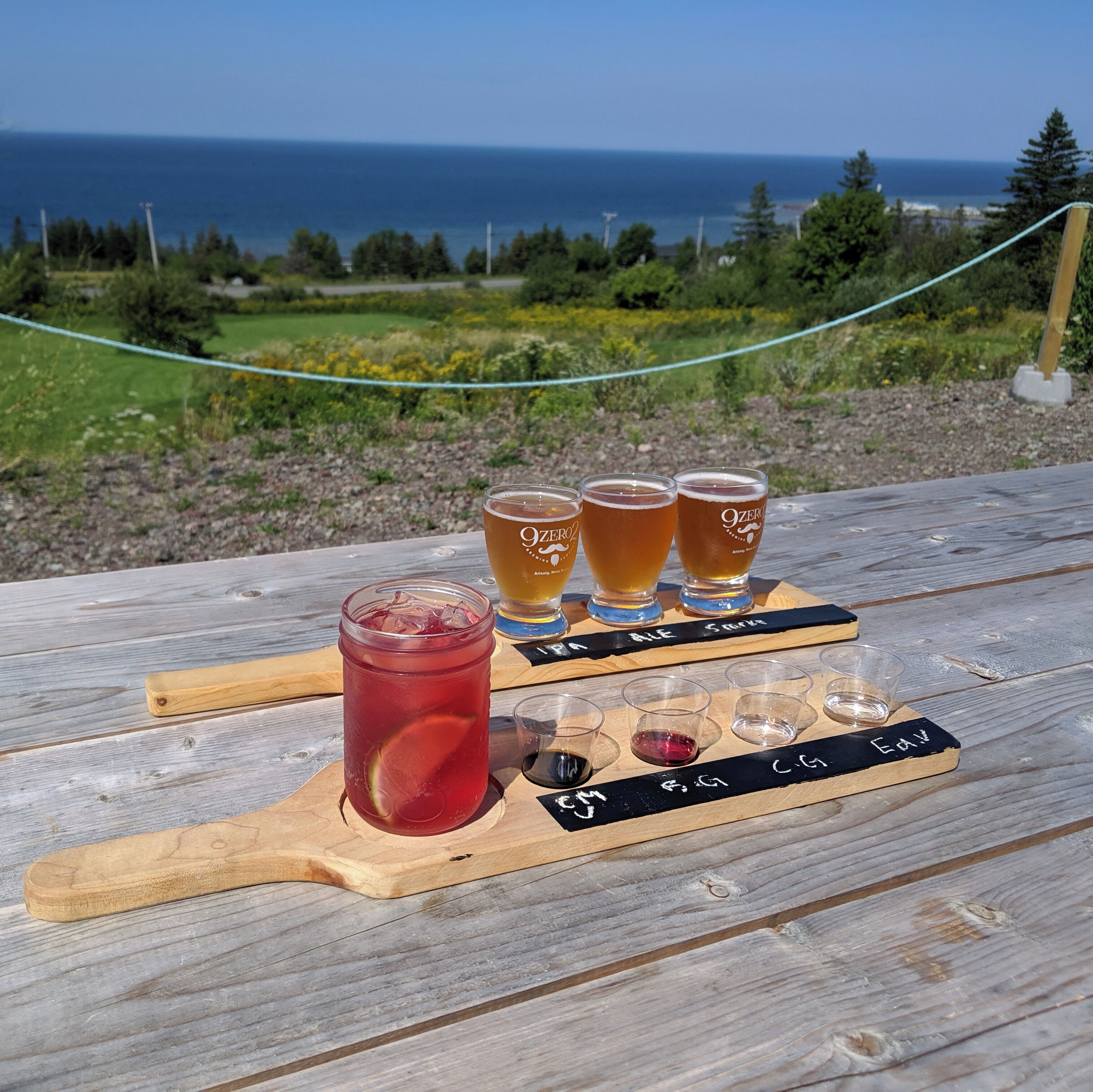 A wooden picnic table with a glass of red drink and four glasses of beer labeled '9ZERO' on a wooden board.