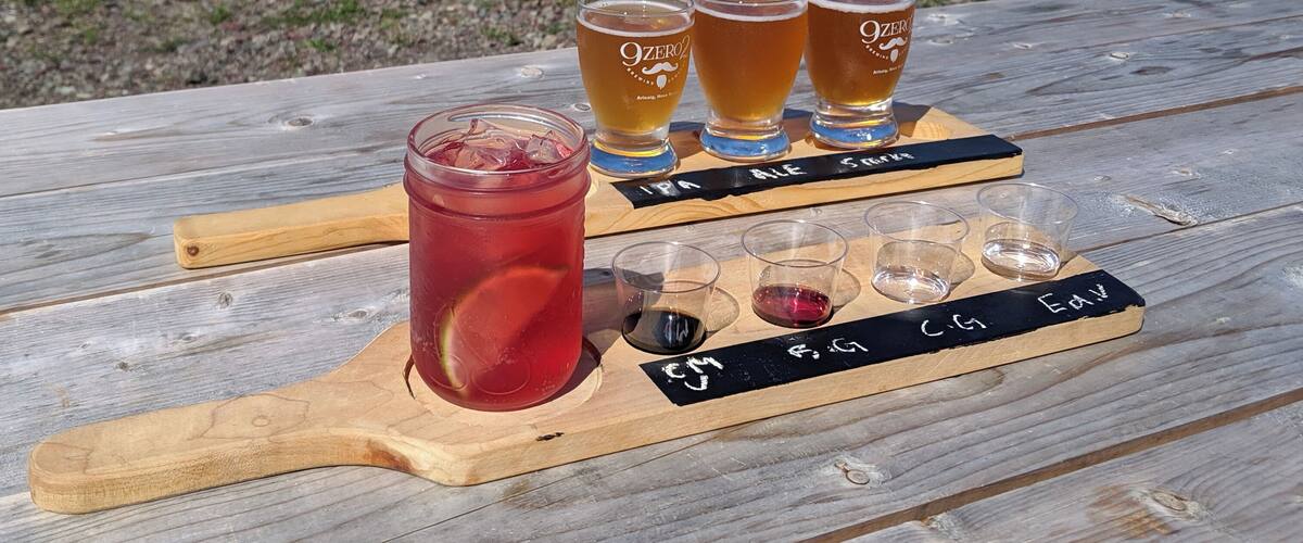 A wooden picnic table with a glass of red drink and four glasses of beer labeled '9ZERO' on a wooden board.