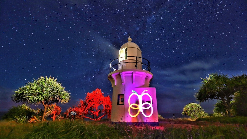 Um farol à noite sob um céu estrelado, iluminado por luzes de neon, rodeado por árvores.