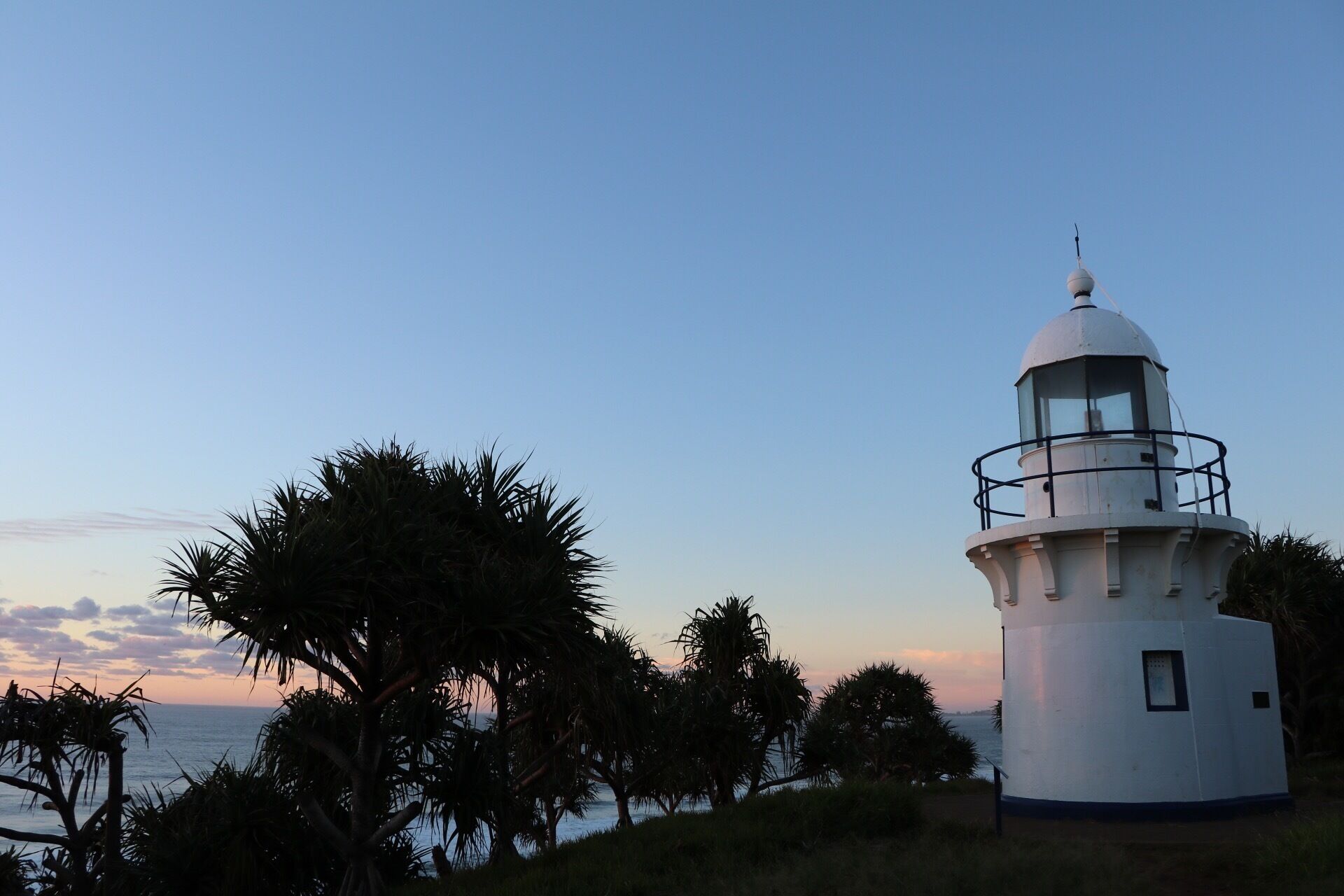 A white lighthouse with a glass lantern room, surrounded by palm trees and overlooking the ocean.