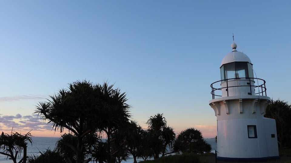 Um farol branco com cúpula de vidro, rodeado por palmeiras e com vista para o oceano.