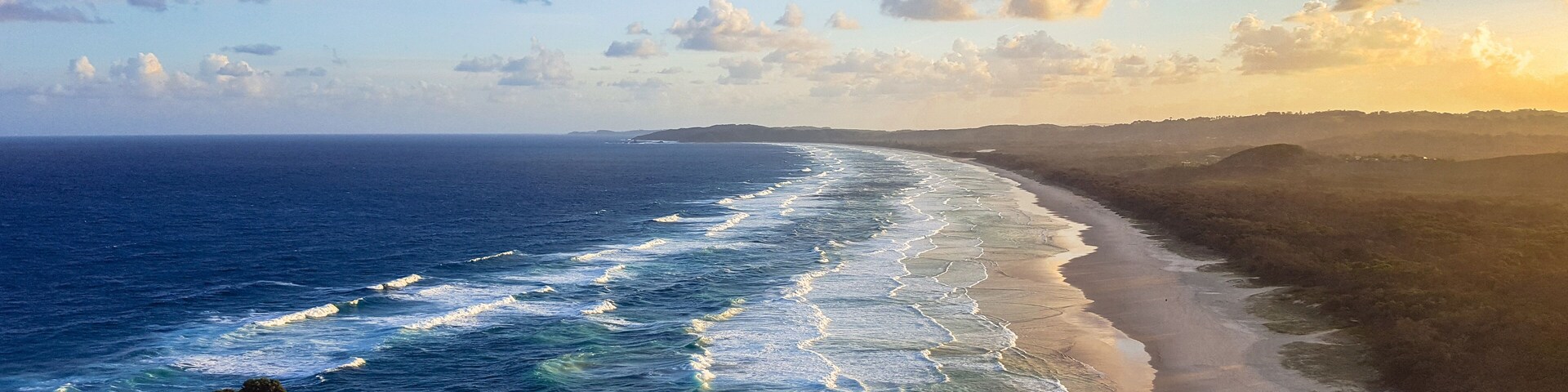 Paisagem costeira com praia arenosa, ondas oceânicas e relevo acidentado coberto por vegetação.