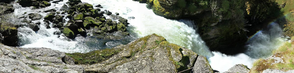 Ein Fluss fließt durch eine bewaldete Schlucht mit felsigem Gelände, im Hintergrund eine Brücke.