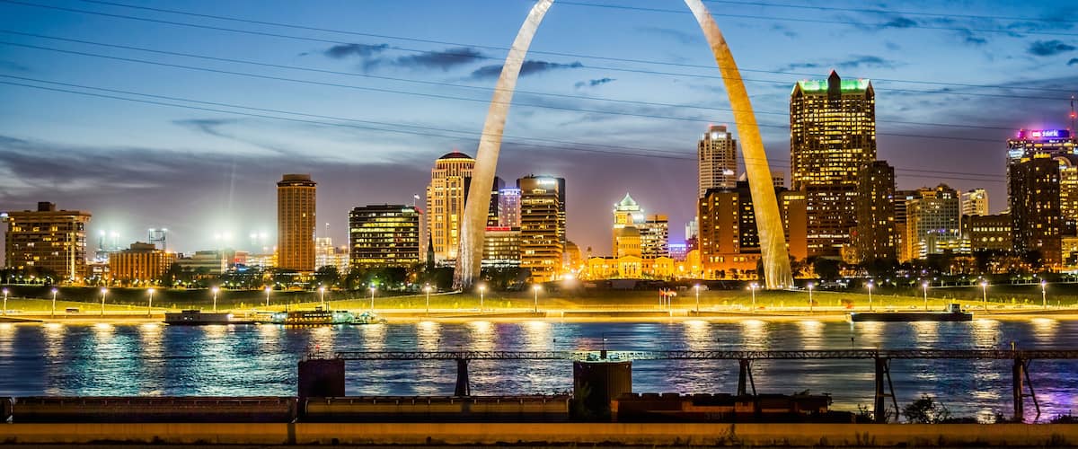 A city skyline with a prominent arch bridge over a river, illuminated buildings, and a clear sky.