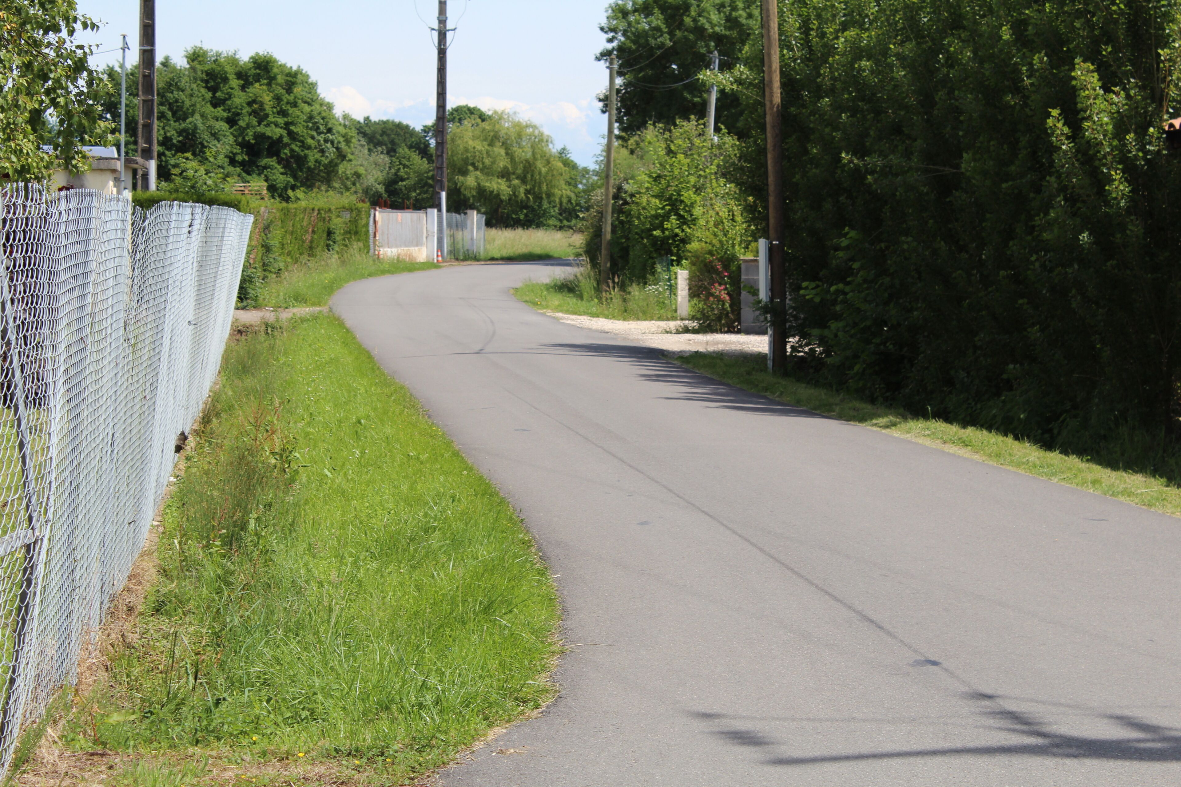 A rural road curving through a green landscape with a chain-link fence on one side and dense trees on the other.