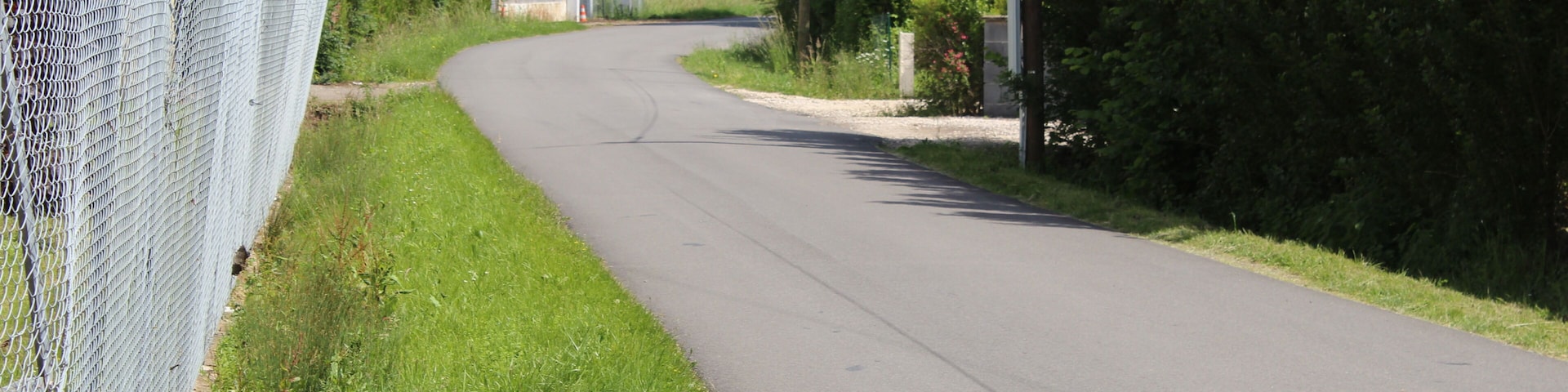 A rural road curving through a green landscape with a chain-link fence on one side and dense trees on the other.