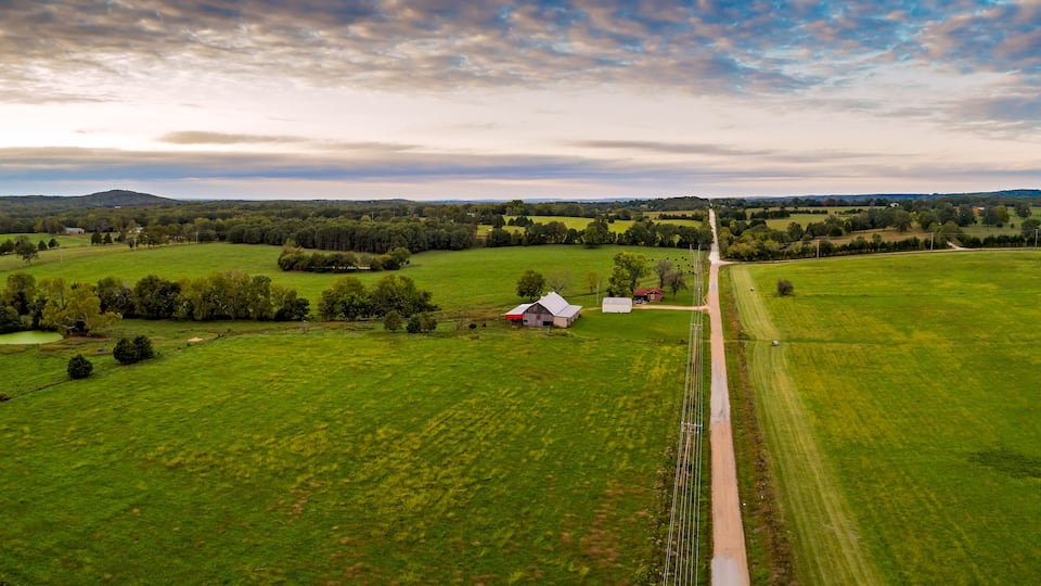 An aerial view of a rural landscape with green fields, a small white barn, and a dirt road.