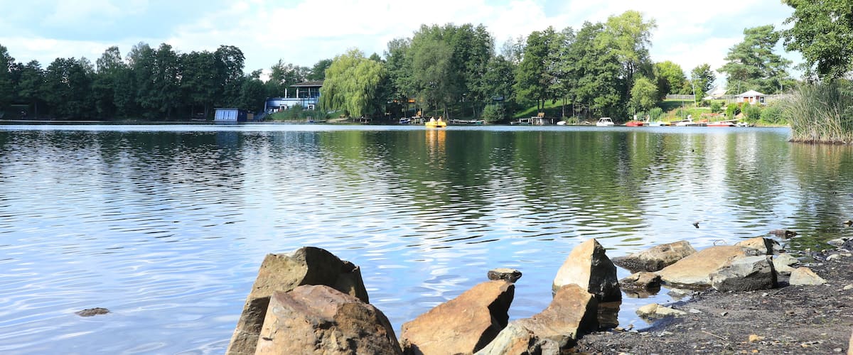 A calm lake with large rocks in the foreground, a small dock, and trees in the background.